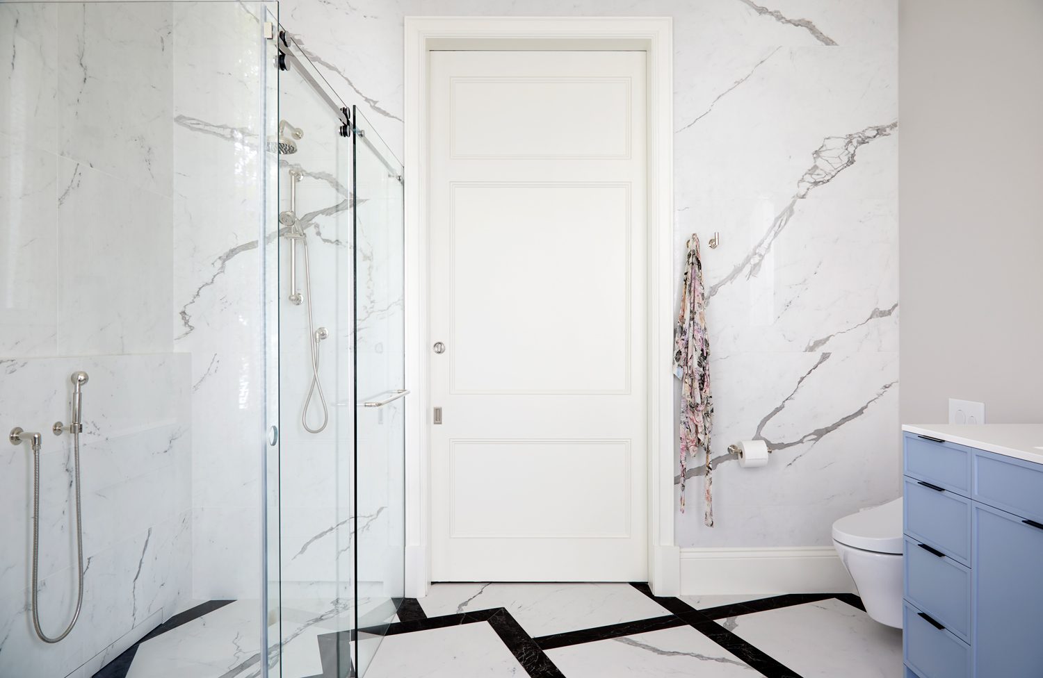 Walk-in shower with frameless glass, marble walls and a low hand shower, beside a blue vanity unit